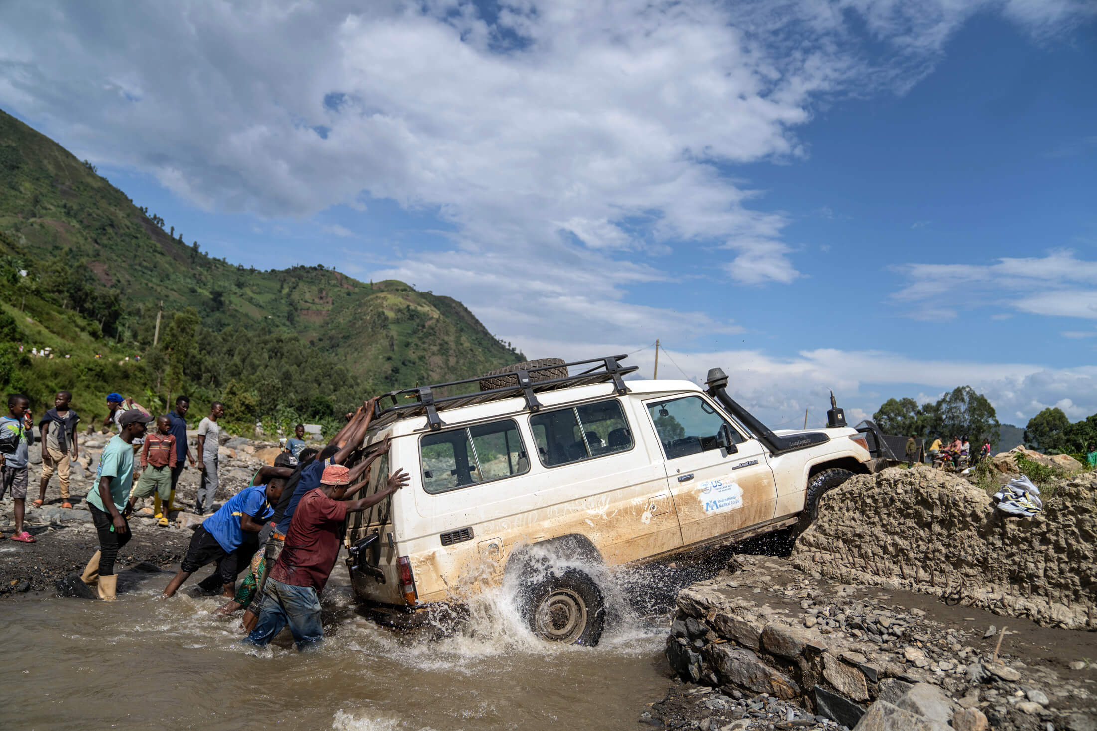 DRC_Moses_IMC_sawasawa_00164 In the hills of South Kivu, local community members help an International Medical Corps team push a mobile medical unit (MMU) across a rocky river, helping our team continue their journey to provide medical services for families displaced from their homes due to violent conflict in the region.